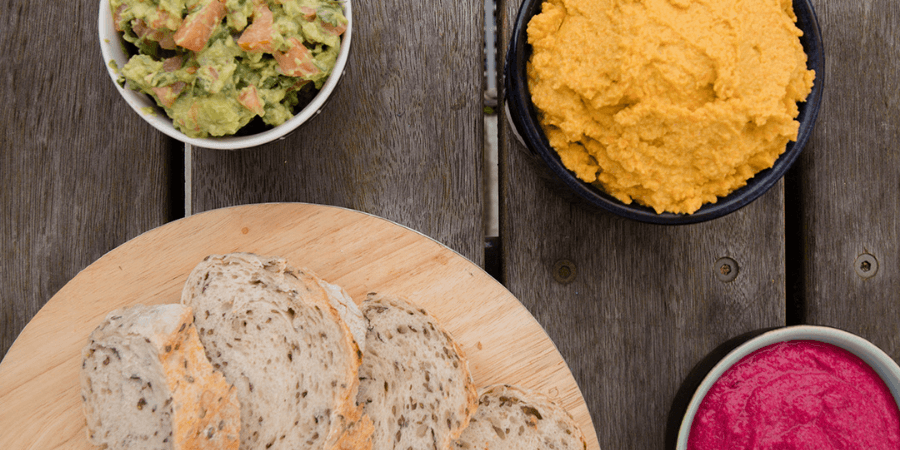 A spread of avocado, beetroot, and carrot and chickpea dips in small bowls served with a plate of bread.
